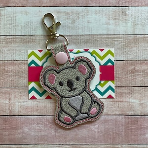 May include: A gray koala keychain with pink ears and a pink button. The keychain is attached to a silver key ring.