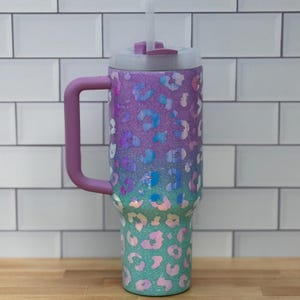 Puede incluir: Un vaso grande y aislado con asa y pajita. El vaso presenta un dise&ntilde;o degradado, que transiciona del morado al turquesa, adornado con un estampado de leopardo. El asa y la tapa son de color lavanda a juego. El vaso est&aacute; dise&ntilde;ado para mantener las bebidas fr&iacute;as.