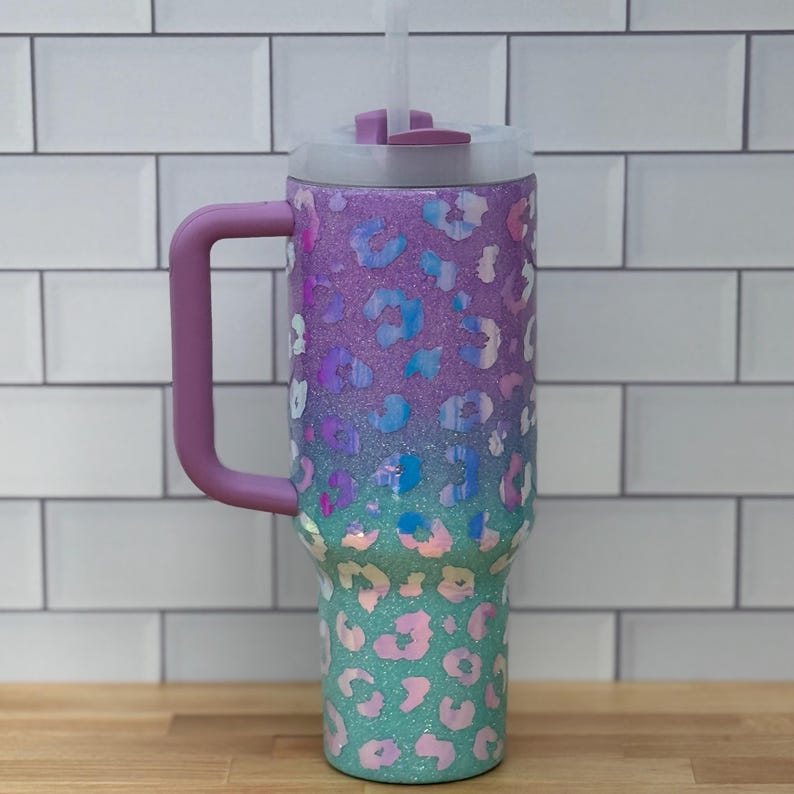 Puede incluir: Un vaso grande y aislado con asa y pajita. El vaso presenta un dise&ntilde;o degradado, que transiciona del morado al turquesa, adornado con un estampado de leopardo. El asa y la tapa son de color lavanda a juego. El vaso est&aacute; dise&ntilde;ado para mantener las bebidas fr&iacute;as.