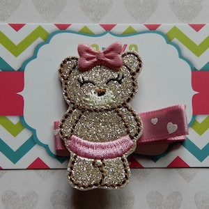May include: A glittery teddy bear hair clip with a pink bow and tutu. The bear is embroidered with brown thread and has closed eyes. The clip is attached to a pink ribbon with white heart accents, set against a colorful chevron background.