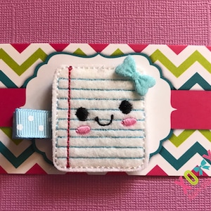 May include: A white felt notepad with blue lines and red lines, a blue bow, and a smiling face. The notepad is on a green and teal chevron background.