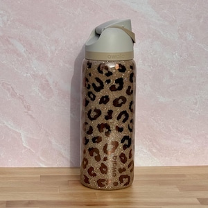 May include: A stainless steel water bottle with a tan and brown leopard print design. The bottle has a gray lid with a handle and the brand name "owala" printed on the bottle.