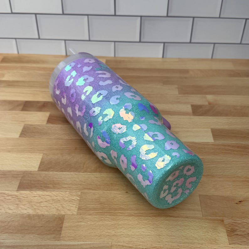 Puede incluir: Un vaso con purpurina y dise&ntilde;o de estampado de leopardo. El vaso hace una transici&oacute;n de un tono p&uacute;rpura en la parte superior a un color turquesa en la parte inferior. Las manchas de leopardo son iridiscentes, agregando un efecto brillante. Incluye tapa transparente y pajita.