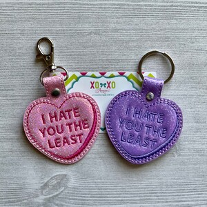 May include: Two heart-shaped keychains with glitter and a stitched border. The pink keychain says "I hate you the least" and the purple keychain says "I hate you the least".