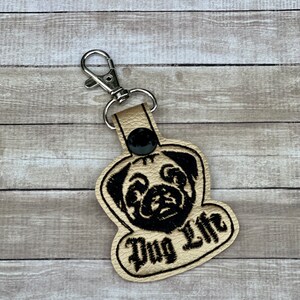 Peut inclure: Un porte-cl&eacute;s en cuir beige avec un visage de carlin brod&eacute; en noir et le texte "Pug Life".