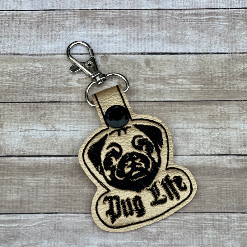 Peut inclure: Un porte-cl&eacute;s en cuir beige avec un visage de carlin brod&eacute; en noir et le texte "Pug Life".