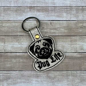 Peut inclure: Porte-cl&eacute;s en paillettes argent&eacute;es avec une silhouette de carlin noire et le texte "Pug Life" en noir.
