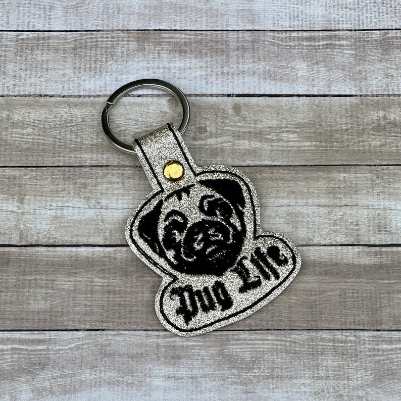 Peut inclure: Porte-cl&eacute;s en paillettes argent&eacute;es avec une silhouette de carlin noire et le texte "Pug Life" en noir.