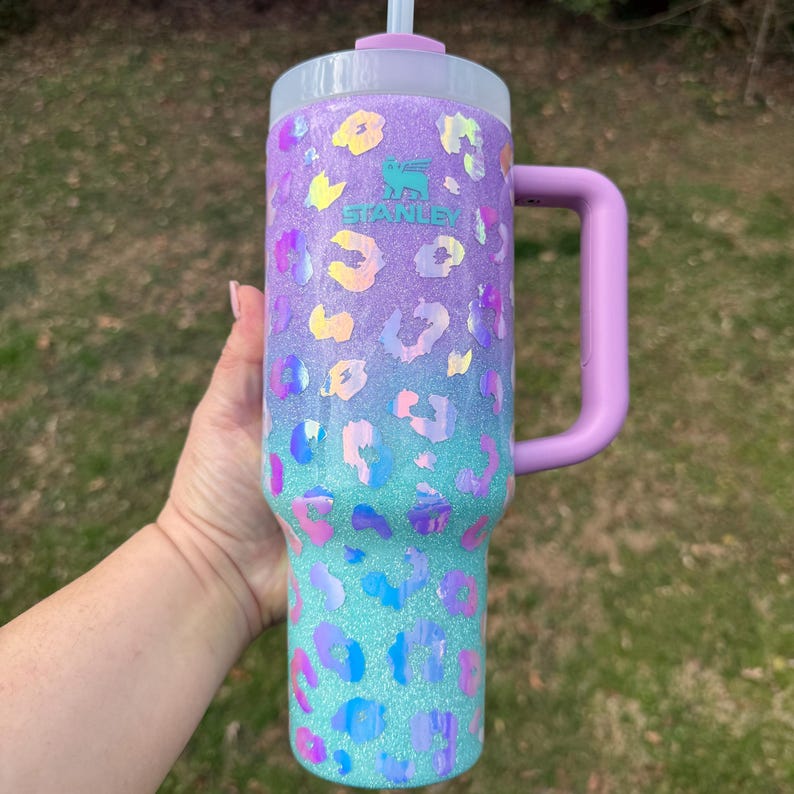 Puede incluir: Un vaso grande y brillante con asa, con un degradado de morado y turquesa con detalles de estampado de leopardo. El vaso tiene un asa y tapa de color morado claro, y el logotipo de Stanley es visible.