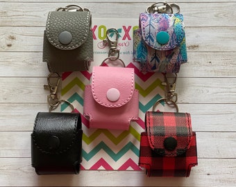 May include: Five different hand sanitizer holders with keychains. The holders are made of different materials and have various patterns, including plaid, feathers, and solid colors. The holders are all attached to silver keychains.