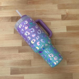 Puede incluir: Un vaso Stanley con asa y tapa moradas. El vaso presenta un dise&ntilde;o degradado, que pasa del morado al turquesa, adornado con manchas de leopardo iridiscentes. Una pajita blanca est&aacute; insertada en la tapa.