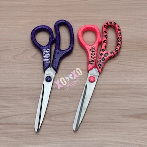 May include: Two pairs of personalized glitter scissors. One pair is purple with the name "Abby" and the other pair is pink with a leopard print and the name "Nicole".
