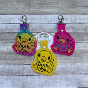 May include: Three yellow duck keychains with the text "Duck You" on them. The keychains are made of felt and have a rainbow, pink, and yellow color scheme.