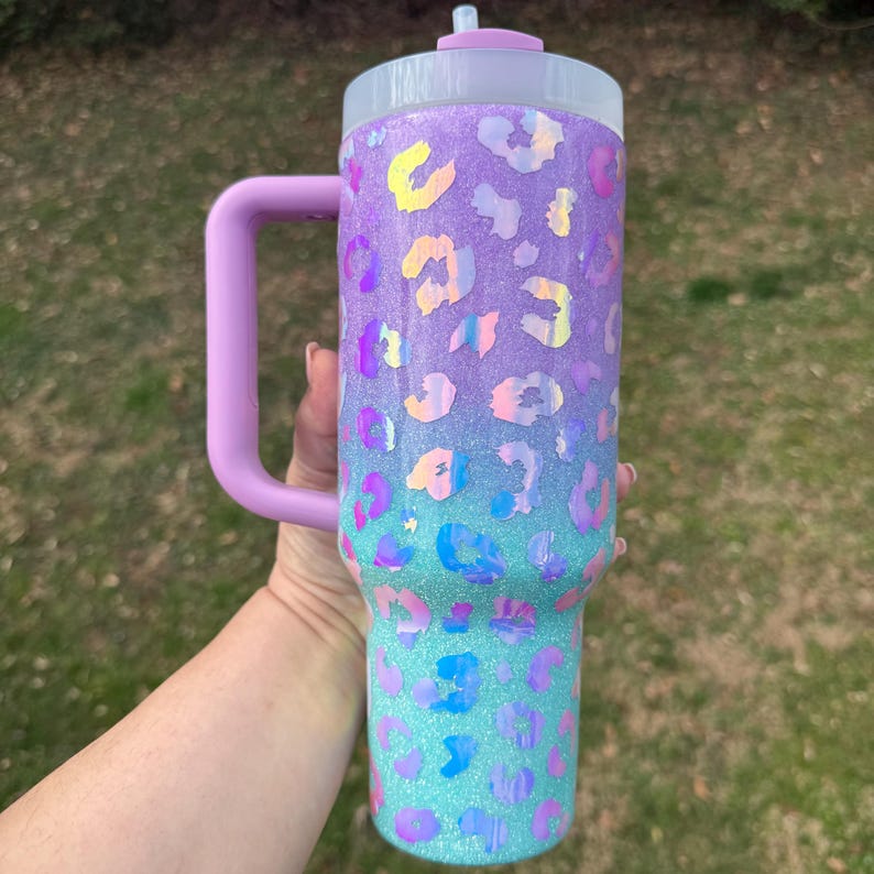 Puede incluir: Un vaso grande y brillante con asa, con un degradado de morado y verde azulado con manchas iridiscentes de estampado de leopardo. La tapa y el asa son de color lavanda liso. El vaso est&aacute; dise&ntilde;ado para bebidas fr&iacute;as.