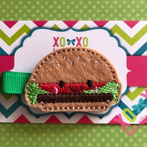 May include: A felt hamburger hair clip with a smiling face, green lettuce, red tomato, and brown patty. The clip is attached to a green ribbon and is displayed on a card with the text "XOXO hair clip couture".