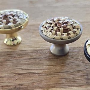 May include: Five different cabinet knobs with woven straw tops. The knobs are in a row on a wooden surface. The knobs are in various colors, including gold, silver, and black.