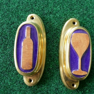 Peut inclure: Deux poignées de meuble en laiton doré sur le thème du vin. L'une représente une bouteille de vin, l'autre un verre à vin, tous deux en couleur bois sur fond violet. Les poignées sont ovales et posées sur une surface verte.