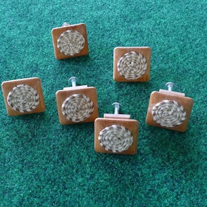 Puede incluir: Seis tiradores cuadrados de madera para armarios con un diseño circular tejido en el centro. Los tiradores son de color marrón claro y tienen un tornillo plateado. El fondo es una superficie verde texturizada.