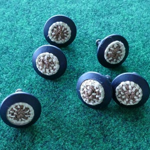 May include: Six dark blue cabinet knobs with a woven design in the centre. The knobs have a circular shape with a white border around the woven centre. They are arranged on a green surface.