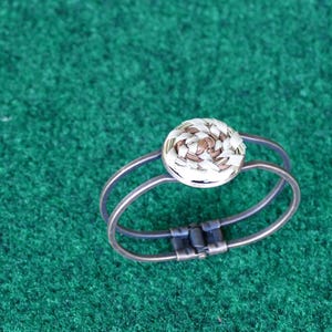 May include: A silver-toned ring with a double band design. The ring features a circular, woven-style centerpiece with white and brown tones. The ring is set against a green background.