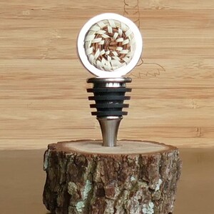 Puede incluir: Un tapón de botella plateado con un diseño tejido marrón y blanco en la parte superior. El tapón está sentado sobre una base de madera.