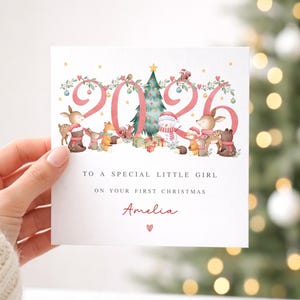 May include: A square Christmas greeting card featuring the year 2026 in large pink font, decorated with watercolor illustrations of woodland animals, a Christmas tree, and gifts. Text reads: TO A SPECIAL LITTLE GIRL ON YOUR FIRST CHRISTMAS Amelia.