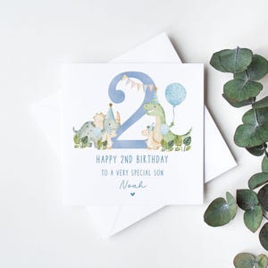 Może przedstawiać: Kartka urodzinowa z dinozaurami w kolorze niebieskim i zielonym z niebieską cyfrą dwa i tekstem "Happy 2nd Birthday to a very special son Noah".