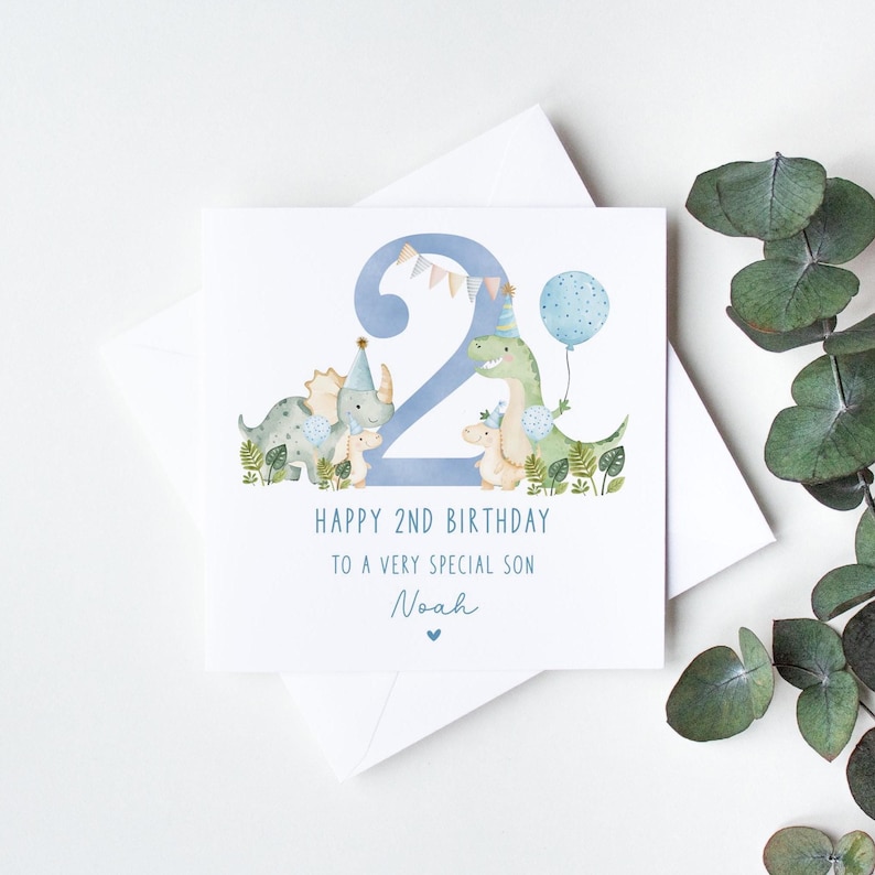 Może przedstawiać: Kartka urodzinowa z dinozaurami w kolorze niebieskim i zielonym z niebieską cyfrą dwa i tekstem "Happy 2nd Birthday to a very special son Noah".