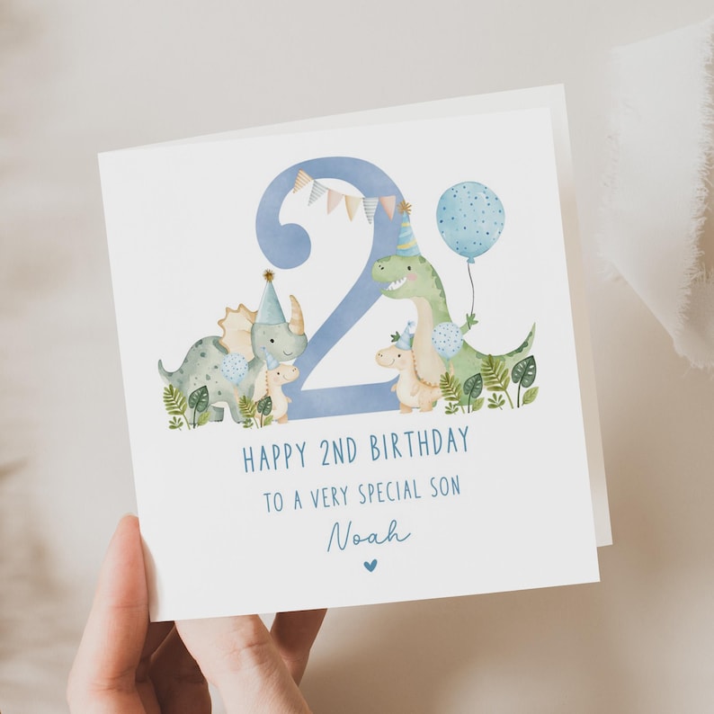 Może przedstawiać: Niebieska i zielona kartka urodzinowa z dużą niebieską cyfrą "2" otoczoną przez dinozaury. Kartka zawiera napis "Happy 2nd Birthday to a very special son Noah".