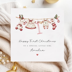 May include: A white card with a watercolor illustration of a clothesline with baby clothes, a stuffed animal, and a Christmas ornament. The text reads "Happy First Christmas to a special little girl Amelia".