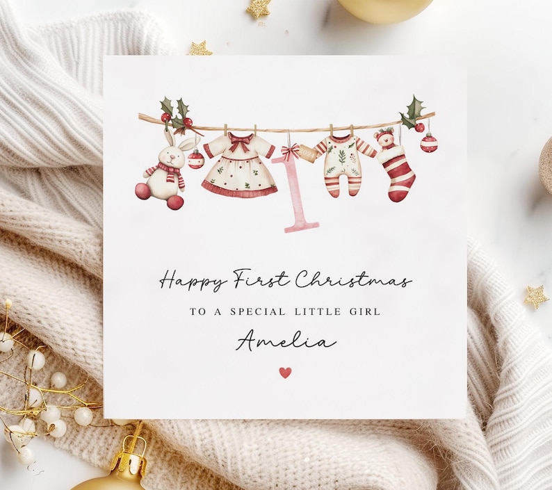 May include: A white card with a watercolor illustration of a clothesline with baby clothes, a stuffed animal, and a Christmas ornament. The text reads "Happy First Christmas to a special little girl Amelia".