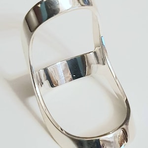May include: A polished silver ring with a distinctive open design. The ring has a rectangular band that curves around the finger, with a horizontal bar across the centre. The reflective surface of the ring creates highlights and shadows.