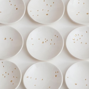 May include: Twelve small, white ceramic plates arranged in a grid. Each plate is decorated with gold star accents. The plates are labeled "VUMBACA WHITE CERAMIC" in the bottom right corner.