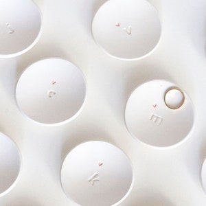 May include: A collection of small, white ceramic ring dishes. Each dish has a letter and a small pink heart. One dish holds a gold ring. The dishes are arranged on a white surface.