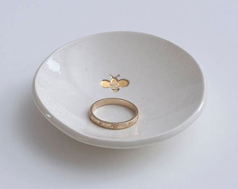 May include: A small, round, white ceramic trinket dish featuring a gold-colored bee illustration at the center. A plain gold-colored metal band ring with a subtle engraved pattern rests inside the dish on the smooth, glossy surface.