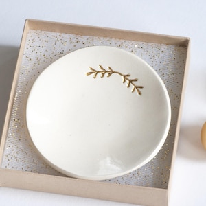 May include: A white ceramic trinket dish with a gold leaf design. The dish is in a box with a white and gold glitter lining.
