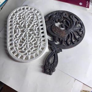 Puede incluir: Dos salvamanteles de hierro fundido. Uno es un salvamanteles blanco de forma ovalada con un intrincado patrón de volutas caladas. El otro es un salvamanteles redondo, de color marrón oscuro y desgastado, con un diseño de gallo.