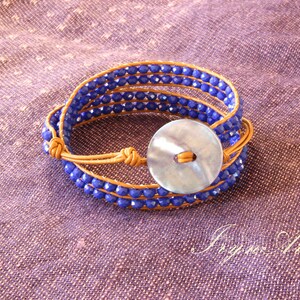 Può includere: Un braccialetto multifilo con perline blu e un avvolgimento in pelle marrone. Il braccialetto ha un grande bottone rotondo e iridescente.