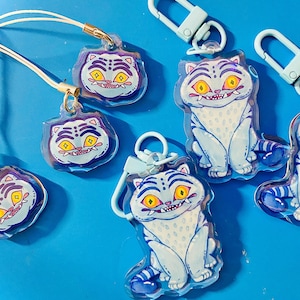 May include: A collection of blue and white cat-themed keychains and charms. The cat design features yellow eyes and a toothy grin. Some charms have a lanyard, while others have a clasp. The background is a solid blue.