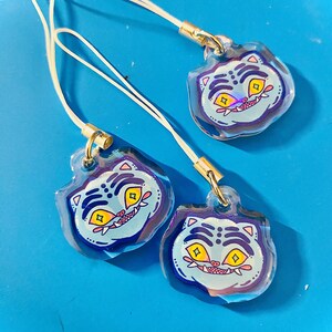 May include: Three clear acrylic charms with a cartoon tiger face design. The tiger has yellow eyes, a red mouth, and blue stripes on a light blue background. Each charm has a white cord and gold-colored metal clasp.