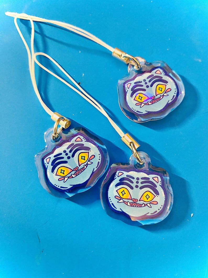 May include: Three clear acrylic charms with a cartoon tiger face design. The tiger has yellow eyes, a red mouth, and blue stripes on a light blue background. Each charm has a white cord and gold-colored metal clasp.