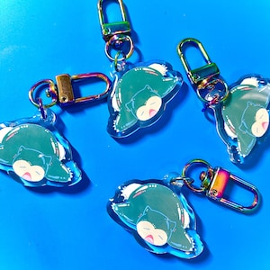 Puede incluir: Cuatro llaveros Snorlax iridiscentes de color verde y azul con cierres de metal plateado y dorado. Cada llavero presenta un Snorlax de dibujos animados durmiendo boca arriba.