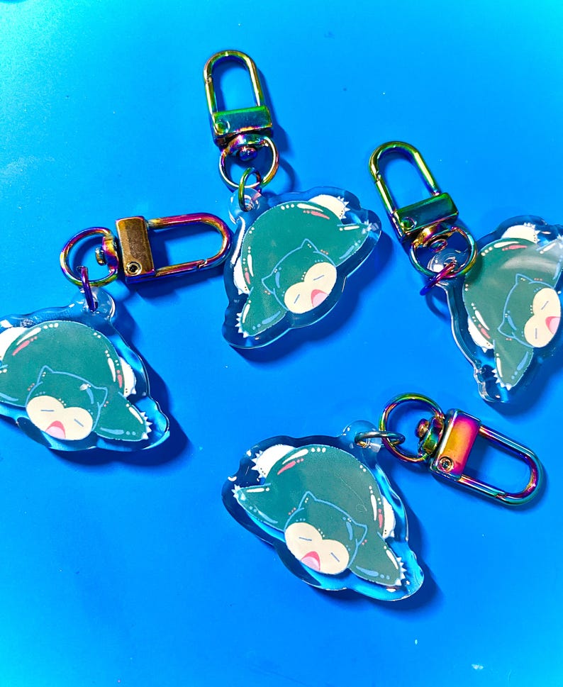 Puede incluir: Cuatro llaveros Snorlax iridiscentes de color verde y azul con cierres de metal plateado y dorado. Cada llavero presenta un Snorlax de dibujos animados durmiendo boca arriba.