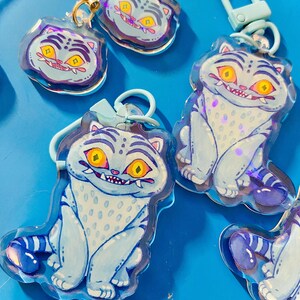 May include: Several cat-shaped keychains with a cartoon design. The keychains are light blue with white spots and stripes, featuring yellow eyes and a toothy grin. They have gold-colored clasps and are arranged on a bright blue surface.