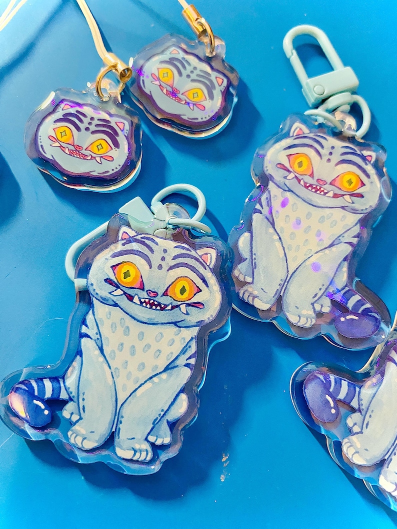 May include: Several cat-shaped keychains with a cartoon design. The keychains are light blue with white spots and stripes, featuring yellow eyes and a toothy grin. They have gold-colored clasps and are arranged on a bright blue surface.