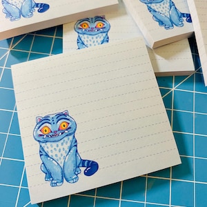 May include: A stack of lined notepads featuring a cartoon cat illustration. The cat is blue with yellow eyes and a striped tail. The notepads have a light blue grid background.