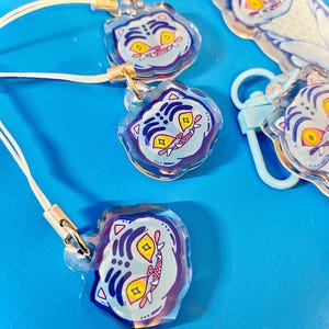 May include: Several clear acrylic charms with a cartoon tiger face design. The tiger face has yellow eyes, a pink mouth, and blue and purple accents. Each charm has a white lanyard and a gold-colored clasp. Some charms are attached to light blue carabiner clips.