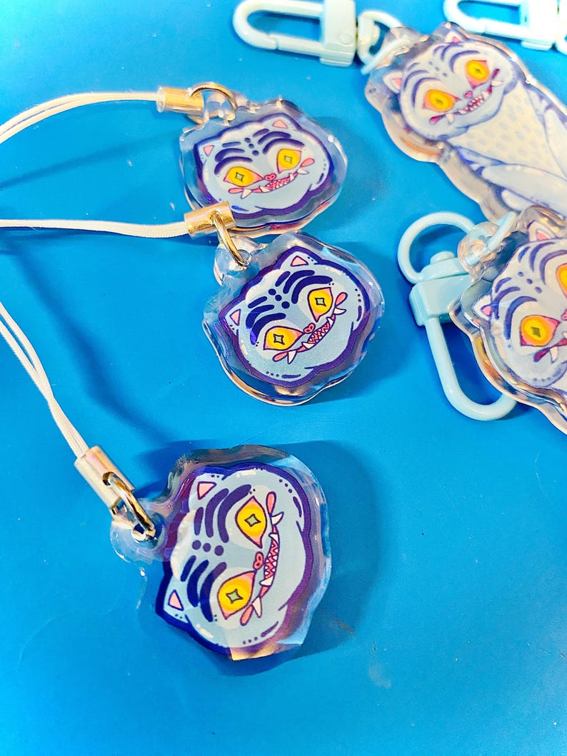 May include: Several clear acrylic charms with a cartoon tiger face design. The tiger face has yellow eyes, a pink mouth, and blue and purple accents. Each charm has a white lanyard and a gold-colored clasp. Some charms are attached to light blue carabiner clips.