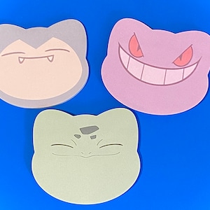 May include: Three cartoon character stickers, each with a different expression. The first sticker is a light brown color with a gray outline and a toothy grin. The second sticker is a light purple color with a gray outline and a wide, toothy grin. The third sticker is a light green color with a gray outline and a closed-mouth smile.
