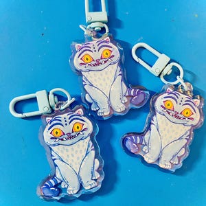 May include: Three cat-shaped keychains with light blue and white coloring, featuring yellow eyes and a purple tail. Each keychain has a light blue clasp. The keychains are on a bright blue background.